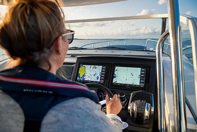 A person with glasses steers a boat, with navigation screens showing maritime maps and a clear waterway ahead.